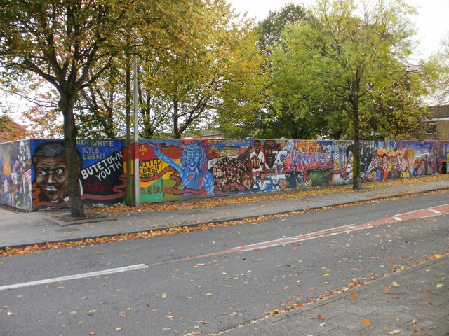 A long colourful mural on a wall beside a tree-lined road in Butetown.