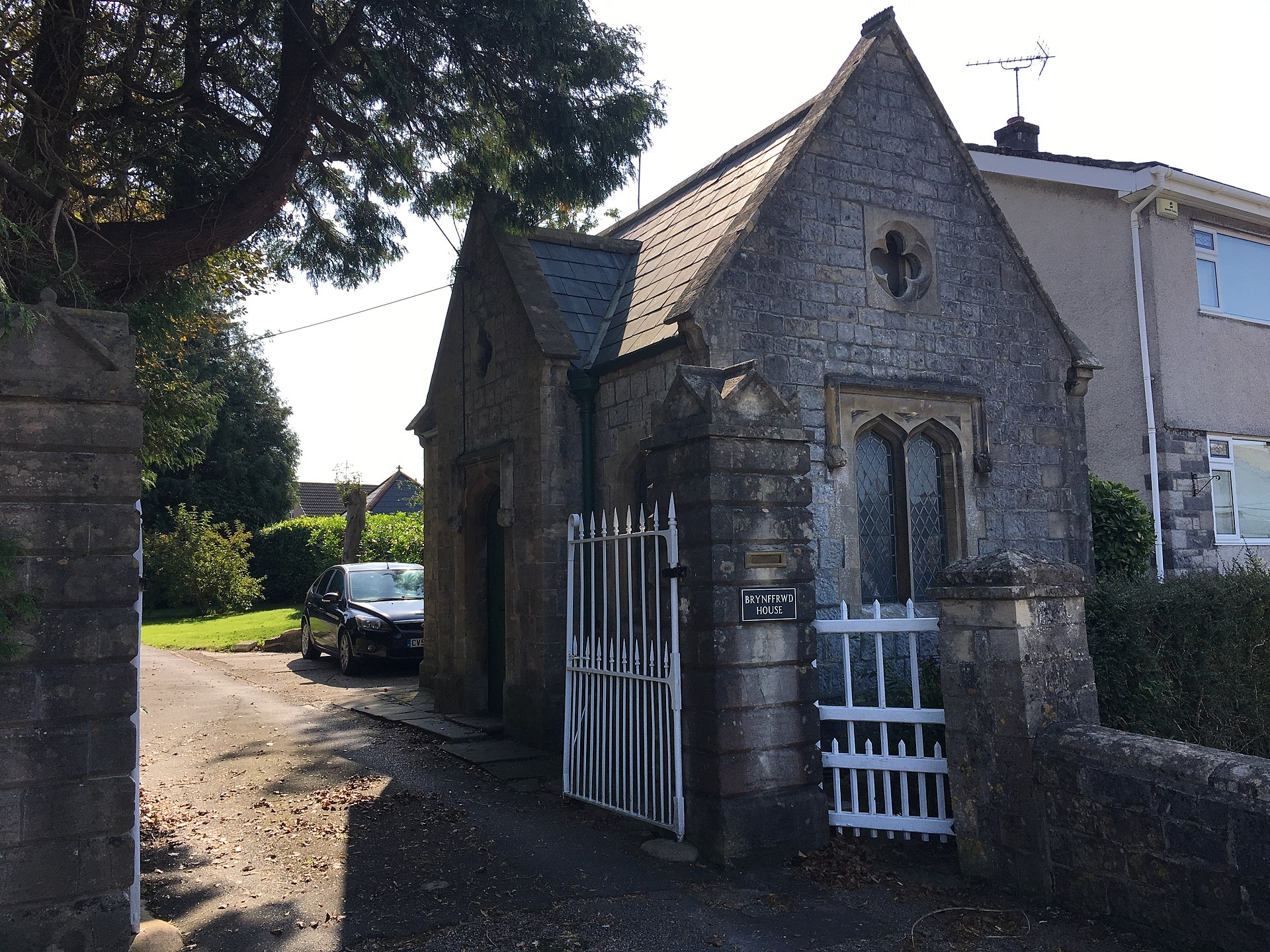 A small grey stone listed building with a pointed gable and iron gates in Coychurch.